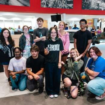 10 high school students stand in a group in front of a new car. One female student is holding the key and smiling.